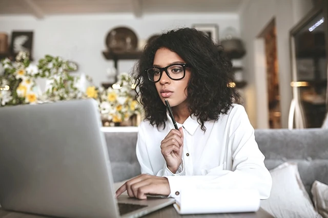 Portrait of lady in glasses thoughtfully looking in laptop with pen in hand